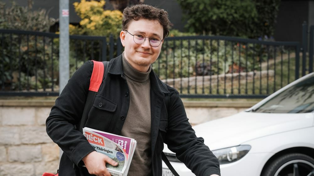 Leon Walter läuft mit einer Zeitung unter dem Arm und einer Umhängetasche die Straße entlang.