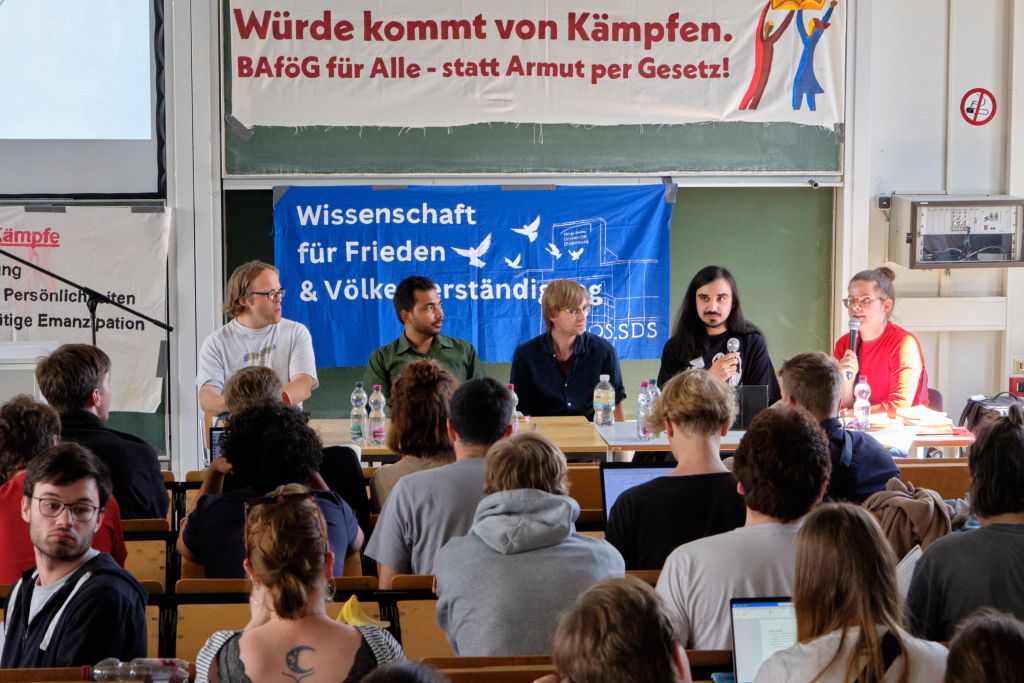 Fünf Personen sitzen in einem Hörsaal an einem Tisch. Vor ihnen sind viele Zuhörer. An der Wand hängen Banner, die ein BAföG für alle fordern.